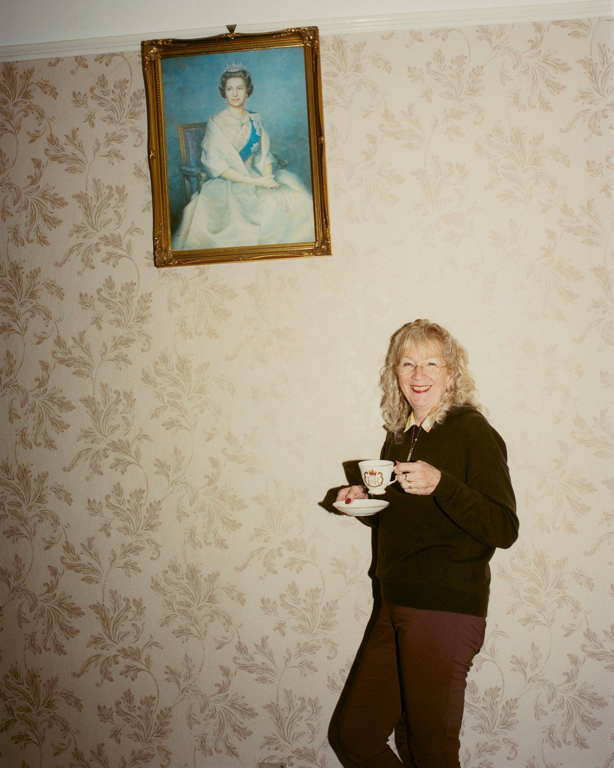 Photo of a woman holding a cup and saucer leaning against a wallpapered wall next to a portrait of Queen Elizabeth II