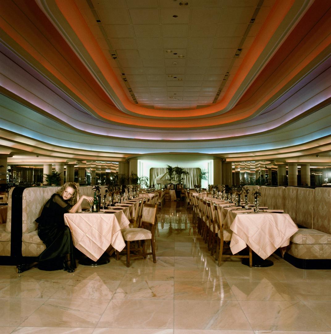 Photograph of the model Twiggy sat on a booth in at the front of a row of tables in a large restaurant space with a ceiling covered in multicoloured curving patterns