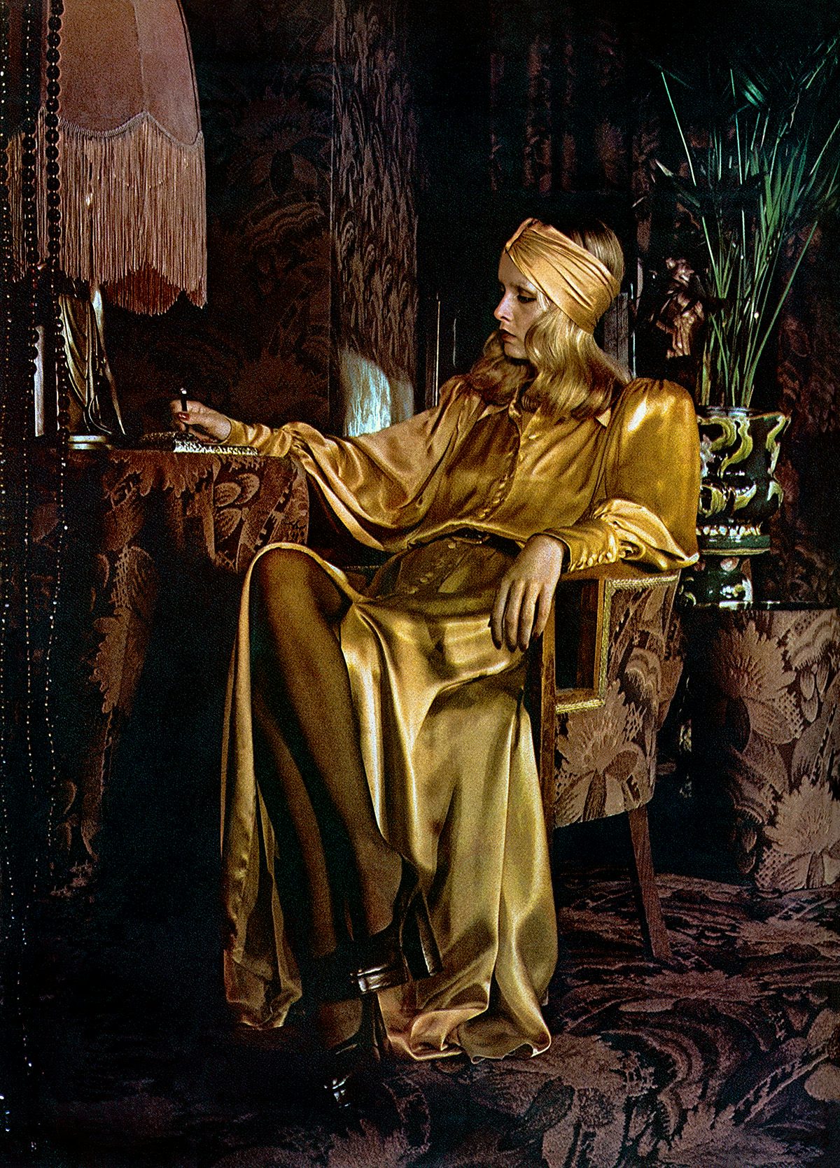 Photograph of the model Twiggy wearing a gold satin dress and headband sat on a chair in an ornate 70s patterned room
