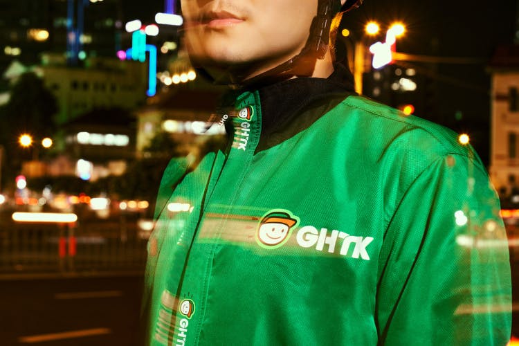 Photo of a person wearing a green jacket featuring the new GHTK branding, including a white uppercase wordmark and a smiling cartoon mascot wearing a cap. Some of the details, like the logo on the jacket, feature motion blur