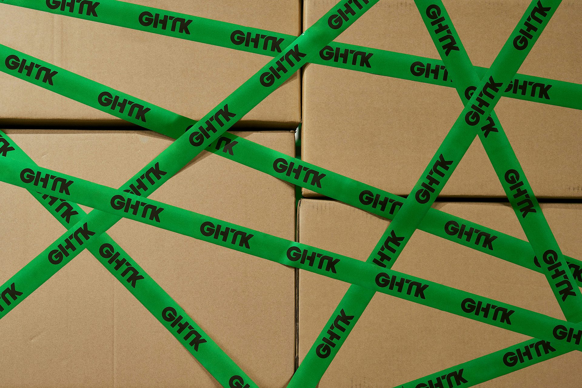 Photo of a stack of cardboard boxes covered in green tape featuring the new GHTK branding, including a black uppercase wordmark featuring cut out details on the letters