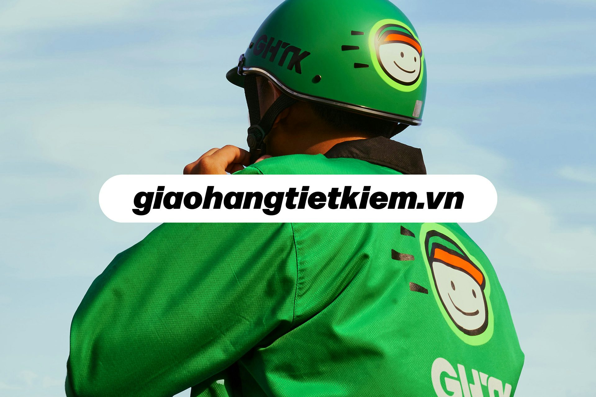 Photo of a person wearing a green jacket and a green helmet, both featuring the new GHTK mascot, a smiling cartoon face wearing a cap. The brand's website address is layered over the photo in black text with a white border