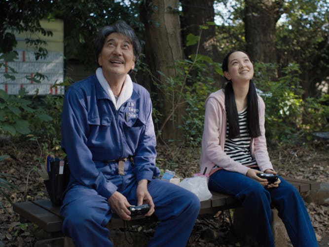 Film still from Perfect Days showing main character Hirayama wearing his Tokyo Toilet uniform sat on a park bench next to his niece, who is wearing long pigtails wearing jeans a pink hoodie