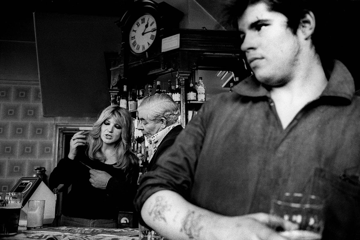 Black and white photograph from Vulcan's Forge showing a person in the foreground holding a pint in a pub, and two people in conversation standing next to a bar in the background