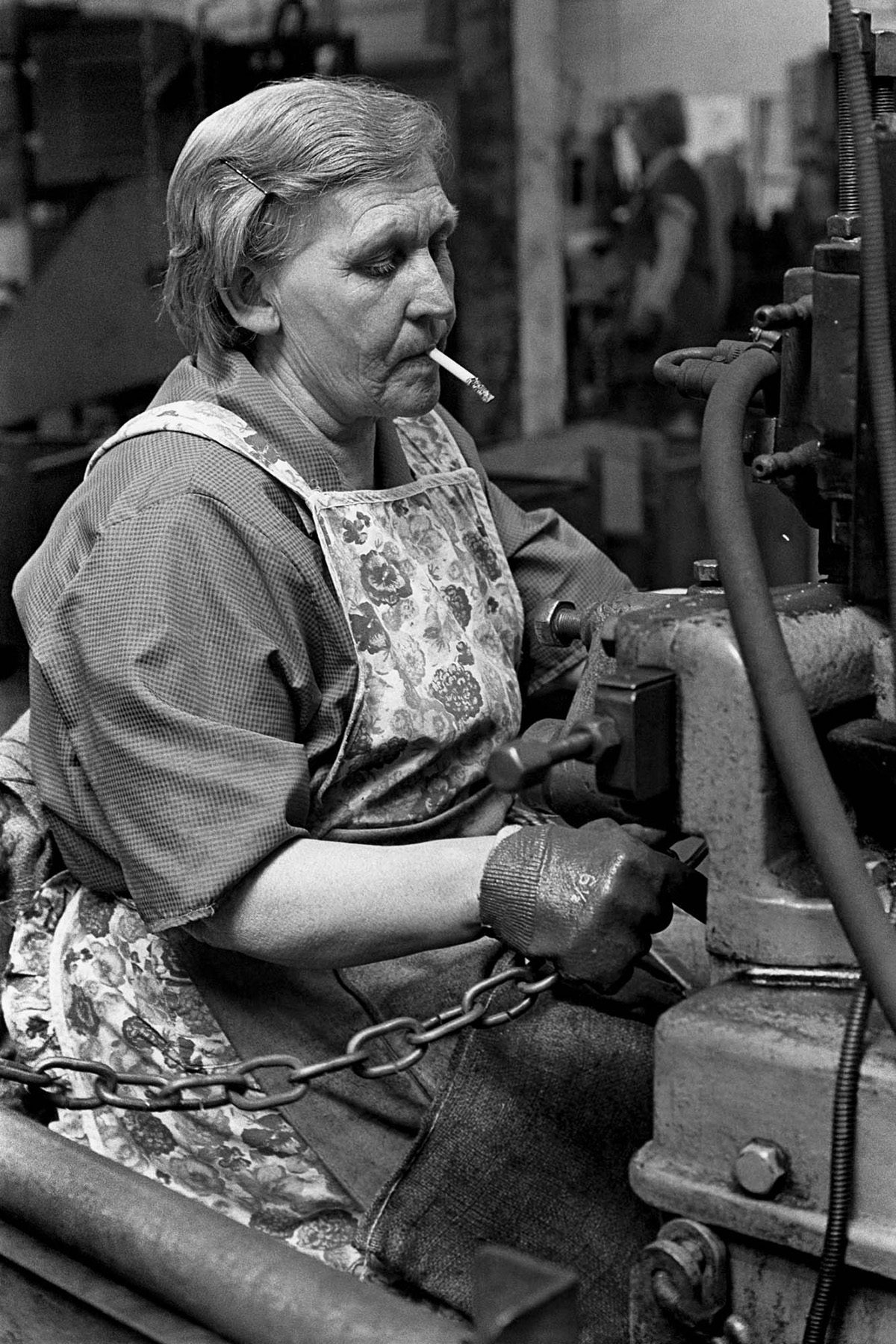 Black and white photograph from Vulcan's Forge showing a person smoking a cigarette while working on making a metal chain