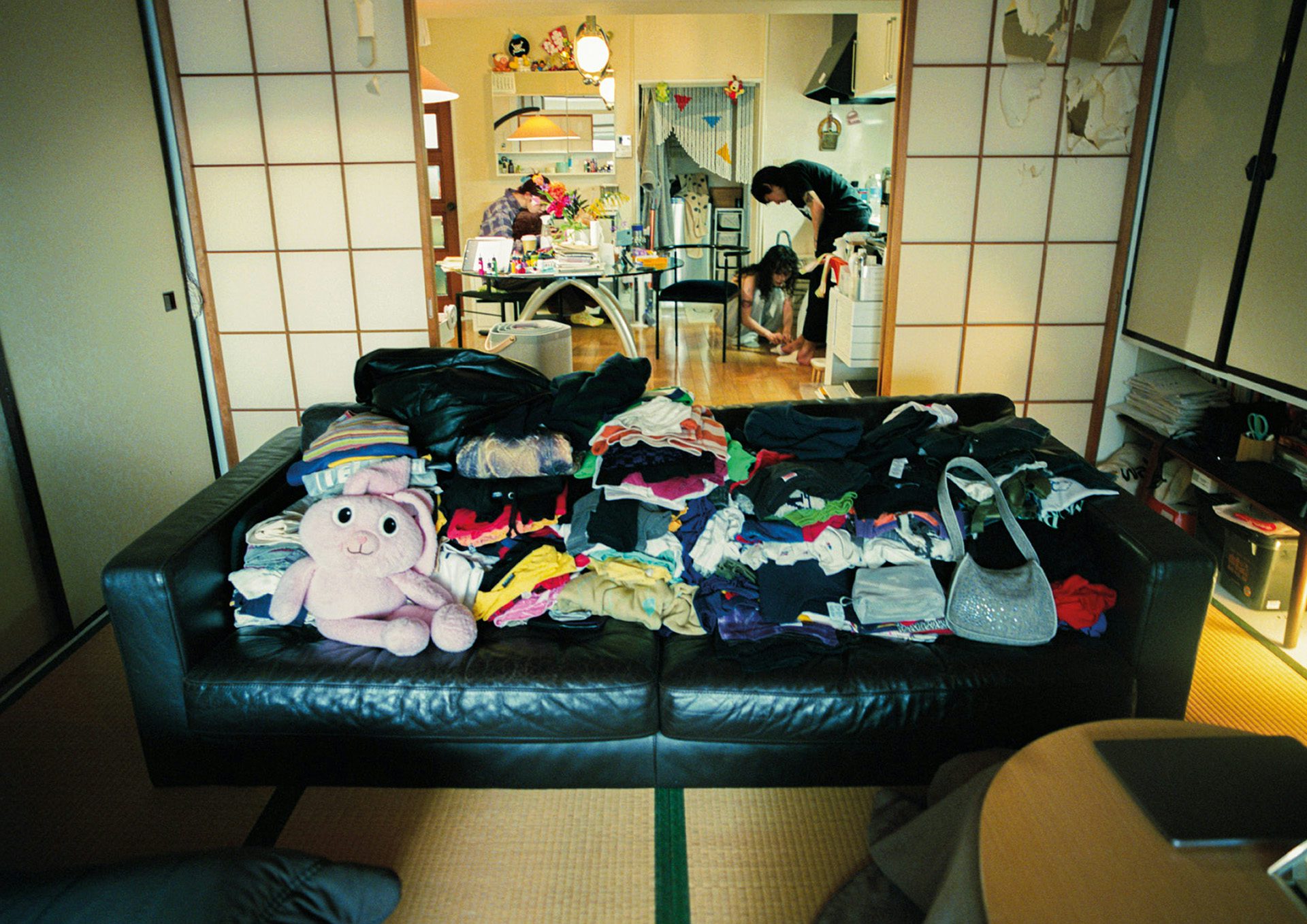 Photograph from Dazed and Ikea's zine showing a dark leather sofa covered in clothing and a pink fluffy toy, with screen doors behind it and three people in the living space in the background