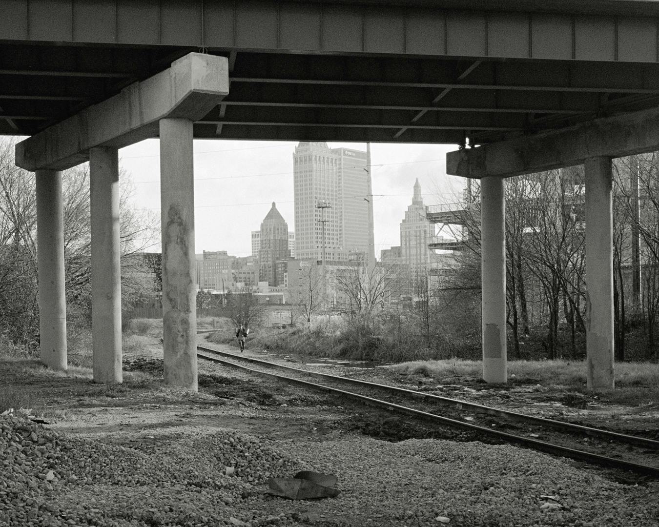 Black and white photograph from Hardtack by Rahim Fortune showing three tall buildings in a city skyline in the background, viewed through a bridge with a trainline running underneath