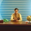 Still image from a Britannia Good Day advert showing a man wearing a yellow long-sleeved shirt and glasses sat behind a desk against a blue background. The lighting and texture of the picture makes it appear like old footage