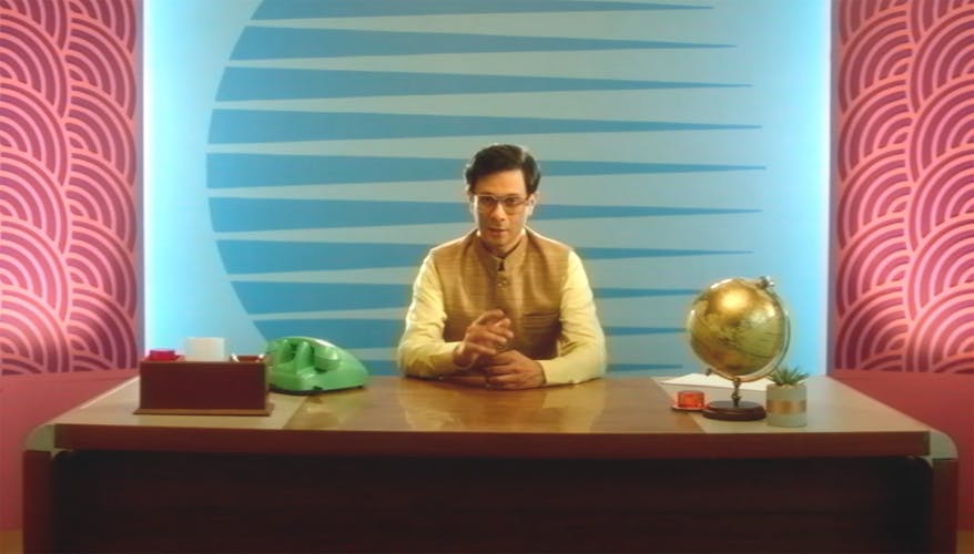 Still image from a Britannia Good Day advert showing a man wearing a yellow long-sleeved shirt and glasses sat behind a desk against a blue background. The lighting and texture of the picture makes it appear like old footage