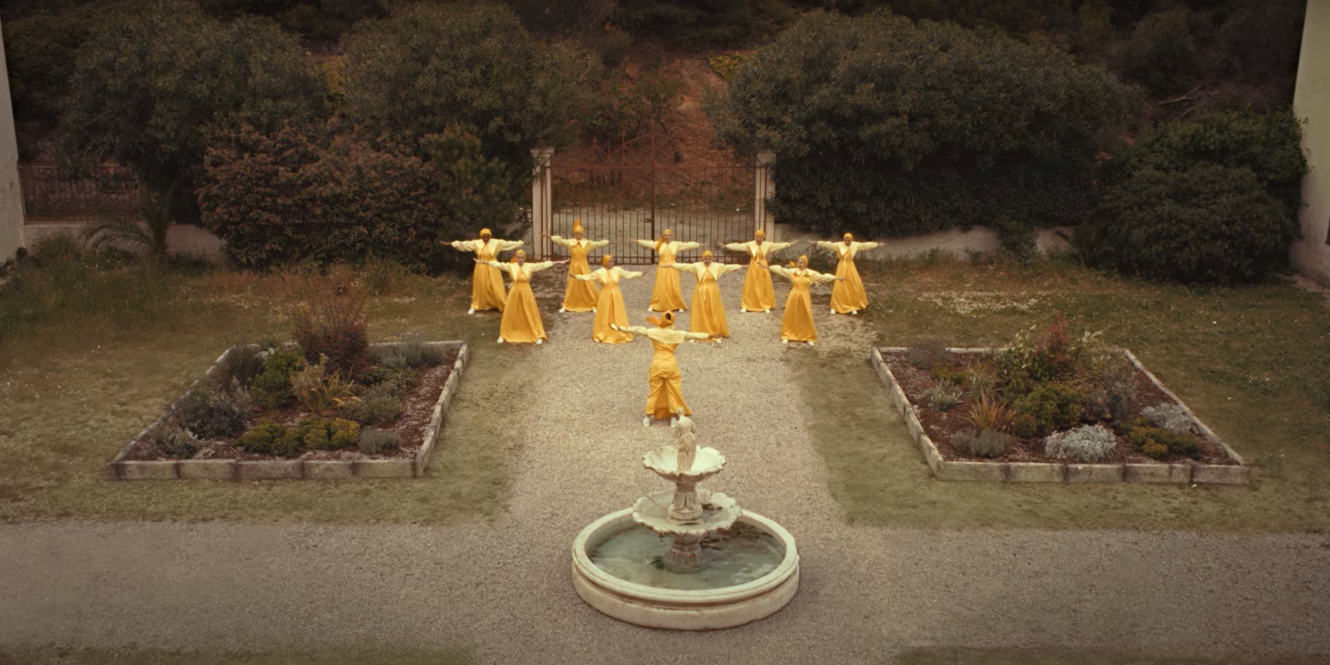 Still from Bumble's campaign advert showing a group of women in long yellow costumes with their arms outstretched in unison while standing in a garden next to a fountain