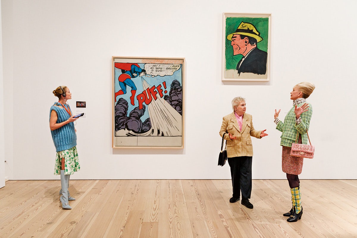 Three people standing in a wooden floored gallery space with two large pop art paintings on the wall