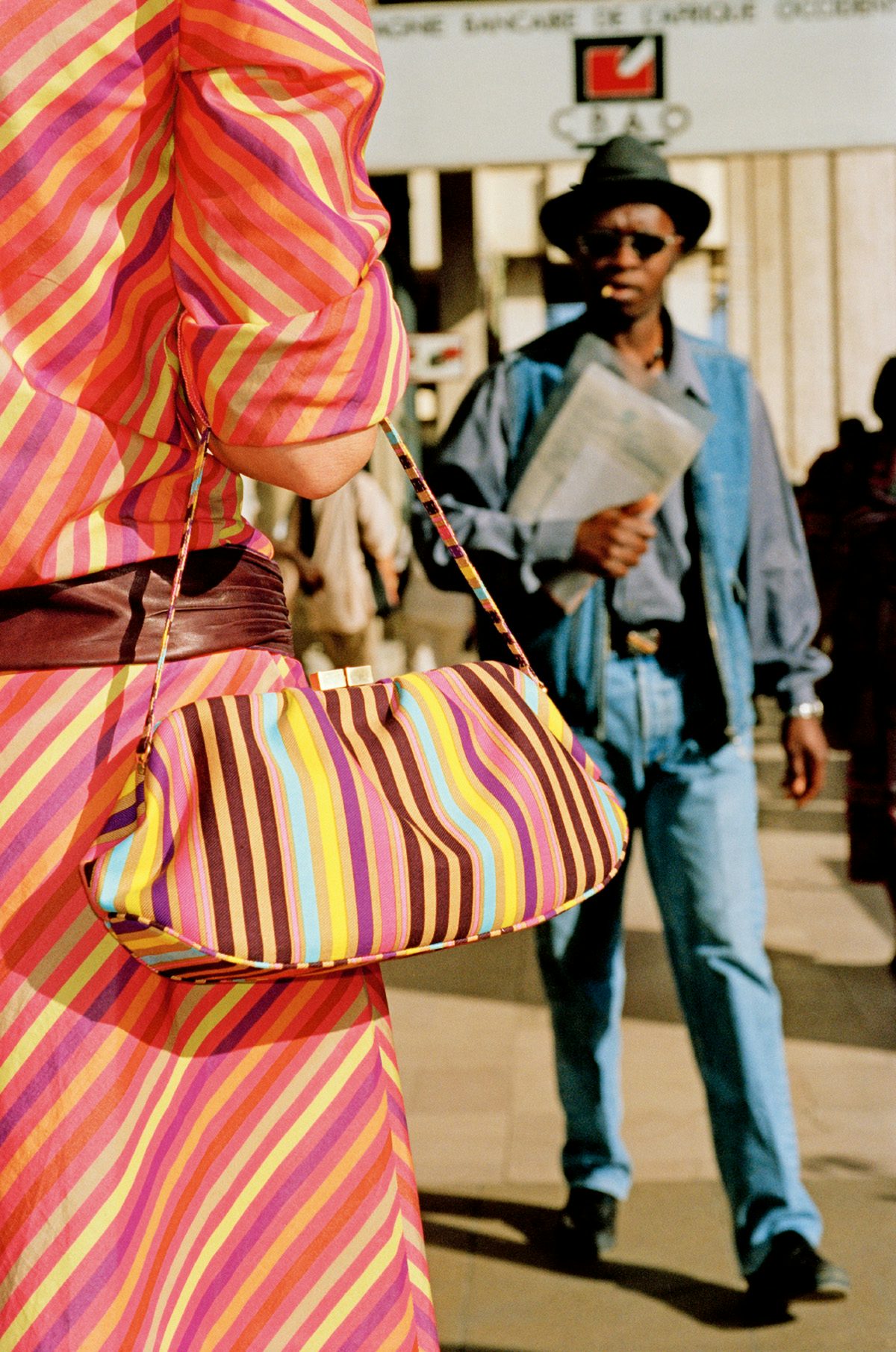 A close up of a person wearing a pink striped dress and bag in the foreground, with a person dressed entirely in denim and a wide-rimmed hat carrying a newspaper in the background