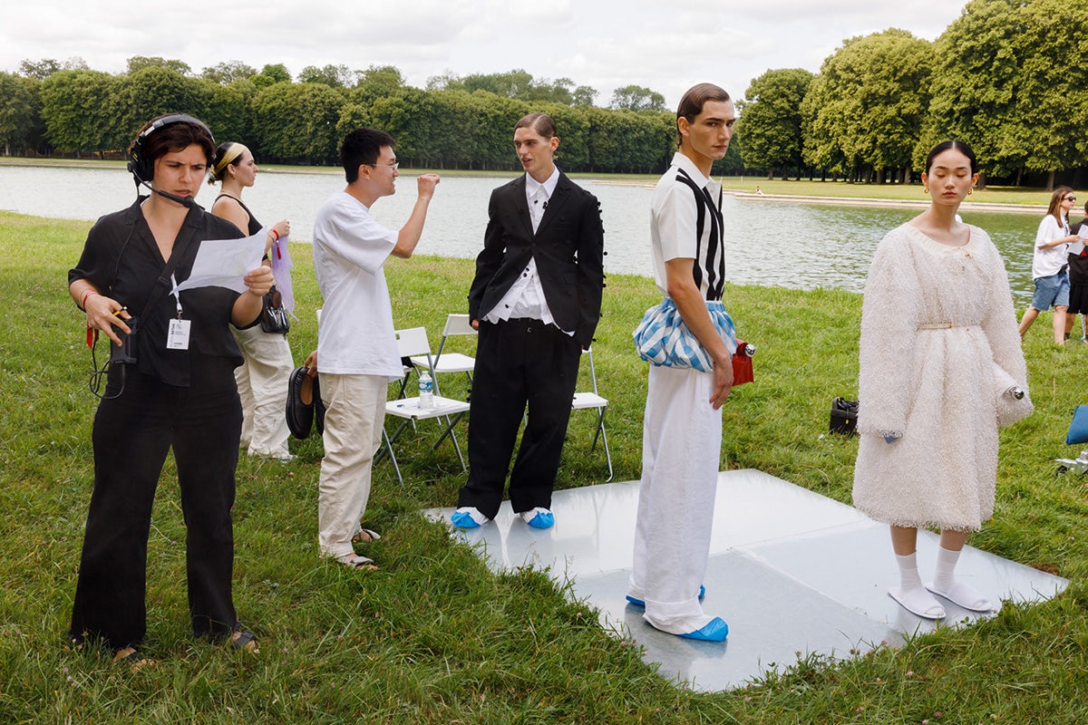 Models on the set of a Jacquemus fashion shoot with an assistant wearing a headset next to them, photographed on grass next to a lake by Martin Parr