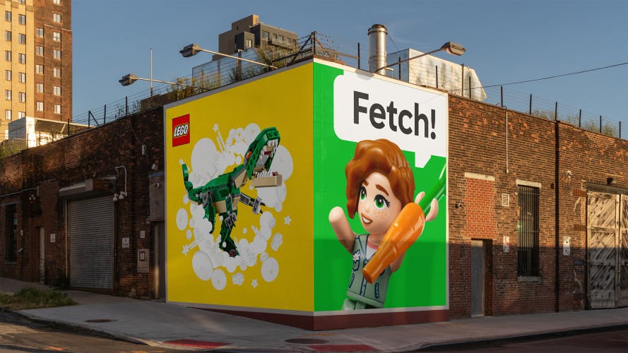 A Lego outdoor advert arranged on two large square outdoor placements wrapped around a corner of a building. The advert shown on the left is a green Lego dinosaur against a yellow background, and the right hand advert shows a Lego character holding a carrot and a speech bubble that reads 'Fetch!'