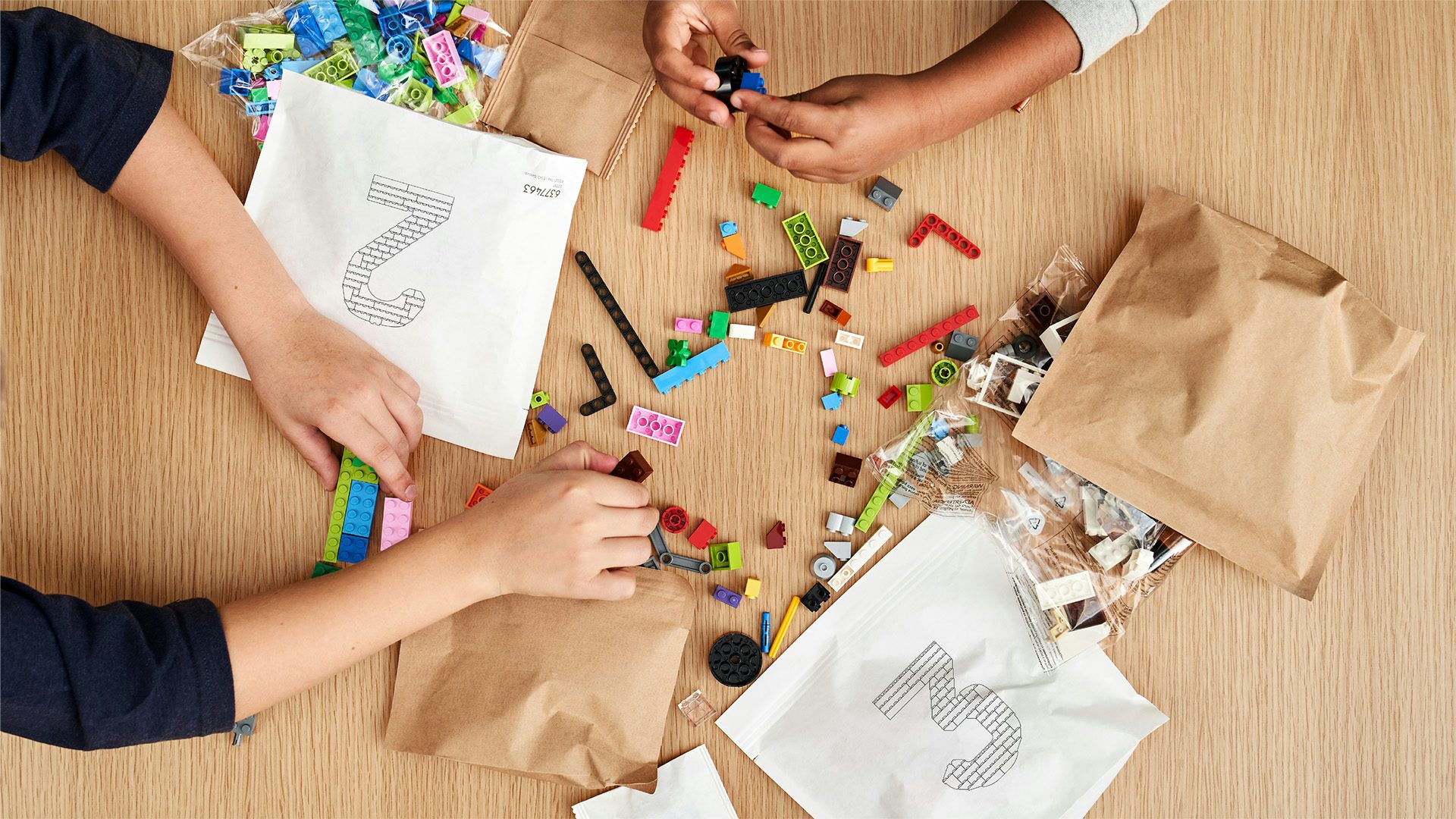 Overhead image the hands of two people playing with Lego pieces, and brown and white paper bags numbered '2' and '3' scattered on a table