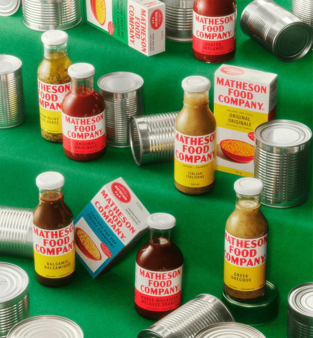 Image shows a green surface covered in bottles of sauce and rectangular boxes labelled with Matheson Food Company branding, along with label-less tin cans