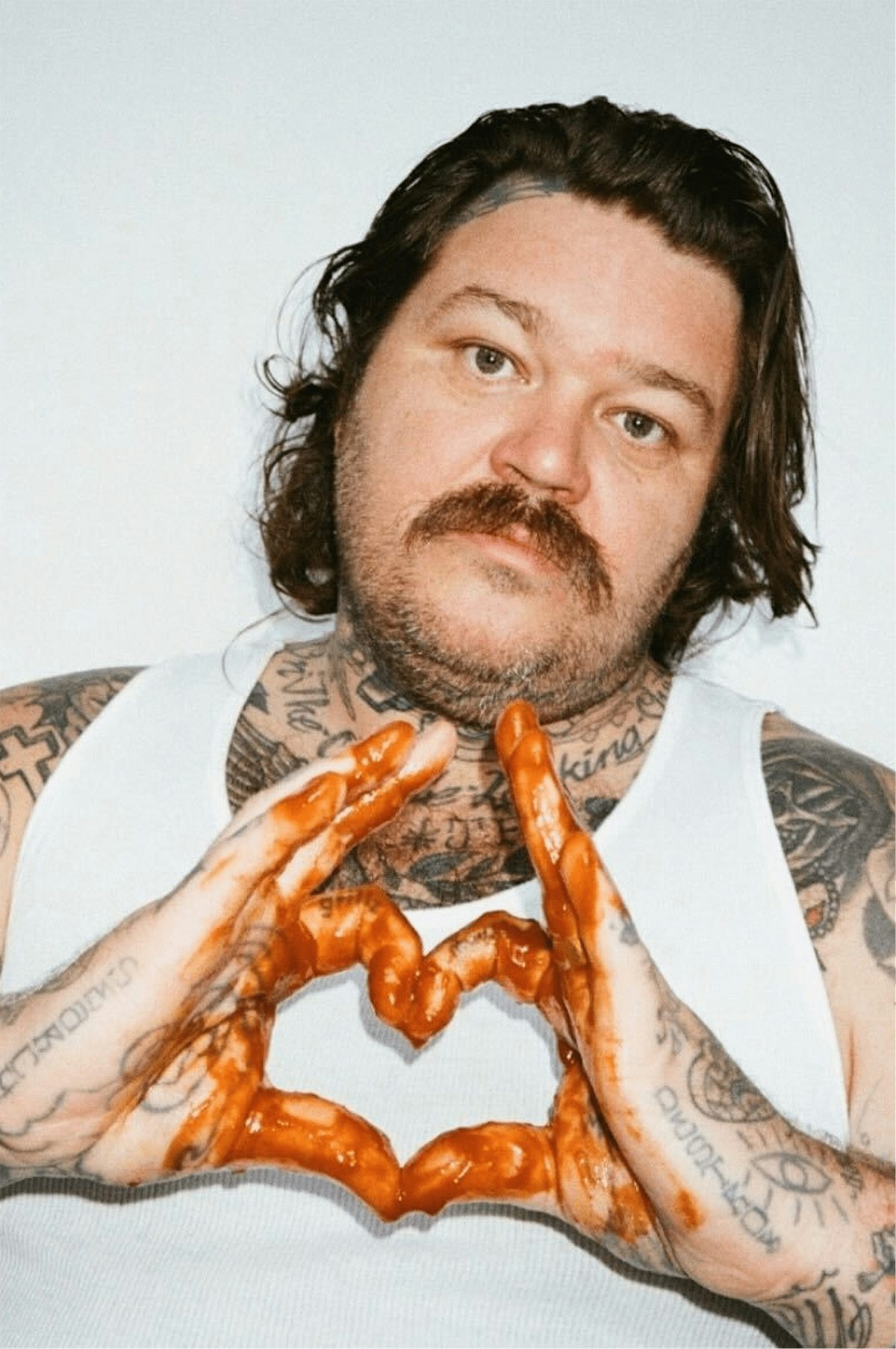 Photograph of chef Matty Matheson wearing a white vest, and joining his sauce-covered hands together in the shape of a heart