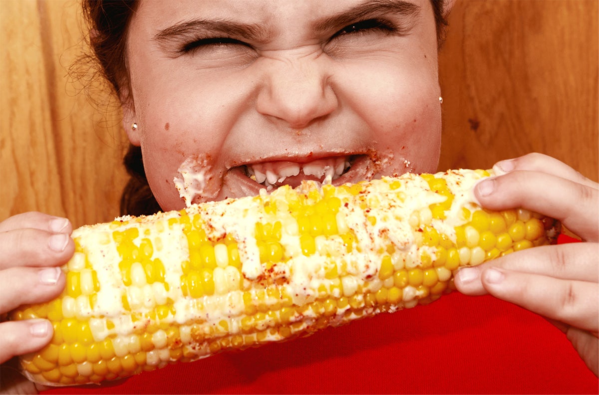 Image shows a child smiling with sauce on their cheeks while holding corncob