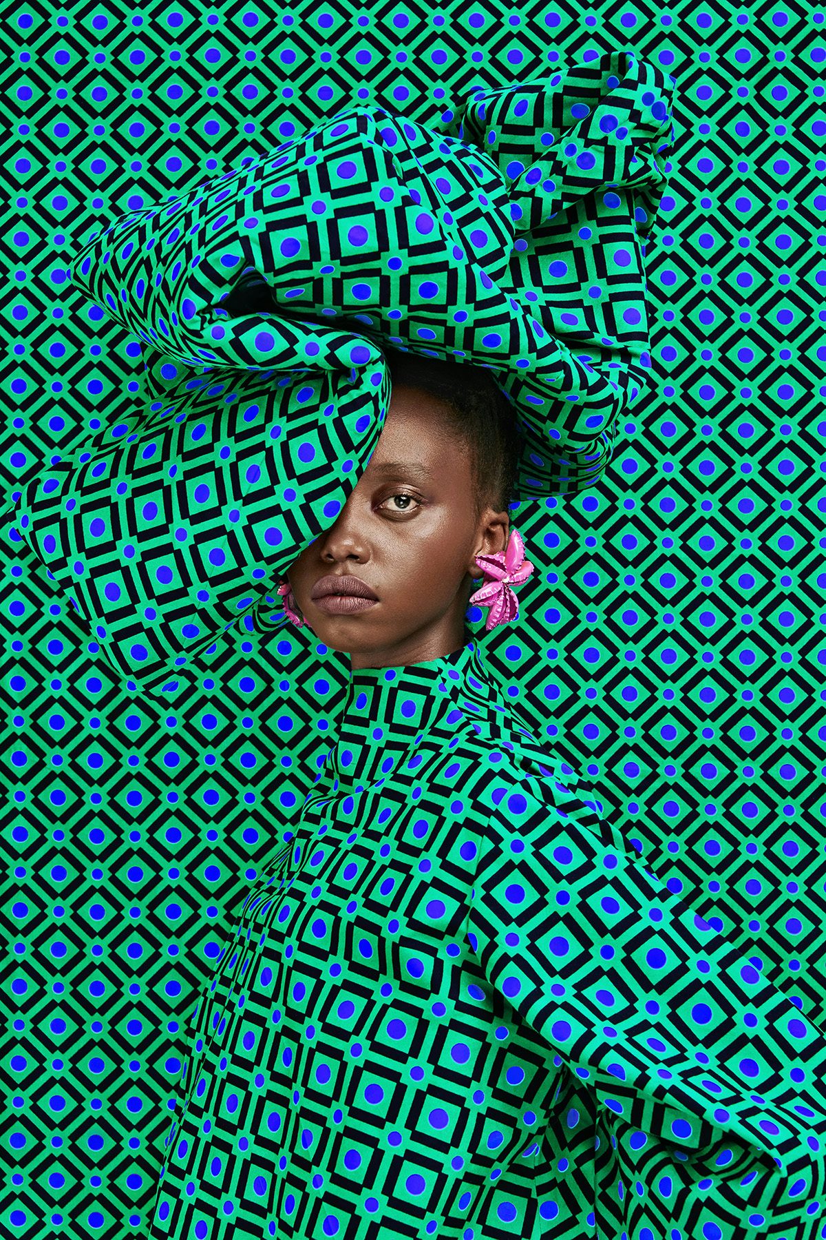 A woman wearing a headdress and garment in a green, blue and black square pattern, which blends into a backdrop in the same pattern