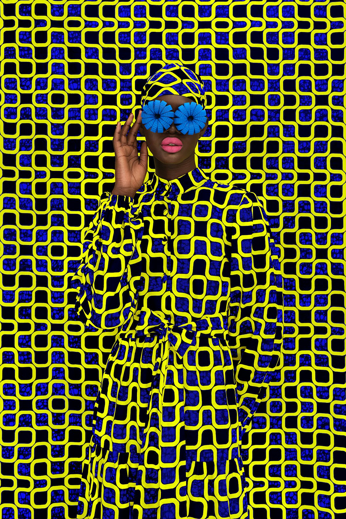 A woman wearing a blue flower-shaped sunglasses, and a headdress and garment in a yellow, blue and black graphic pattern, which blends into a backdrop in the same pattern