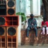 An adult and two young people sat on a wall next to a speaker stack in the street