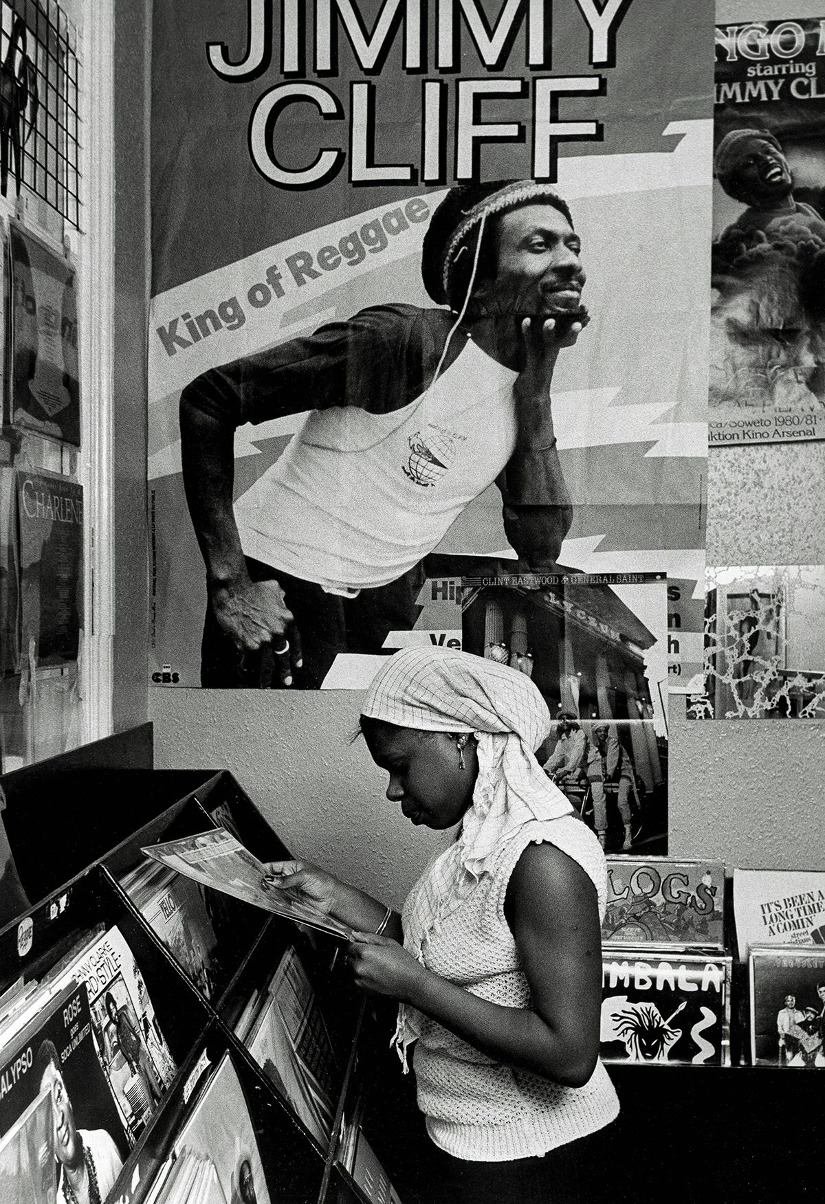 Black and white photo of a person looking at a vinyl record in a store standing beneath a poster of Jimmy Cliff
