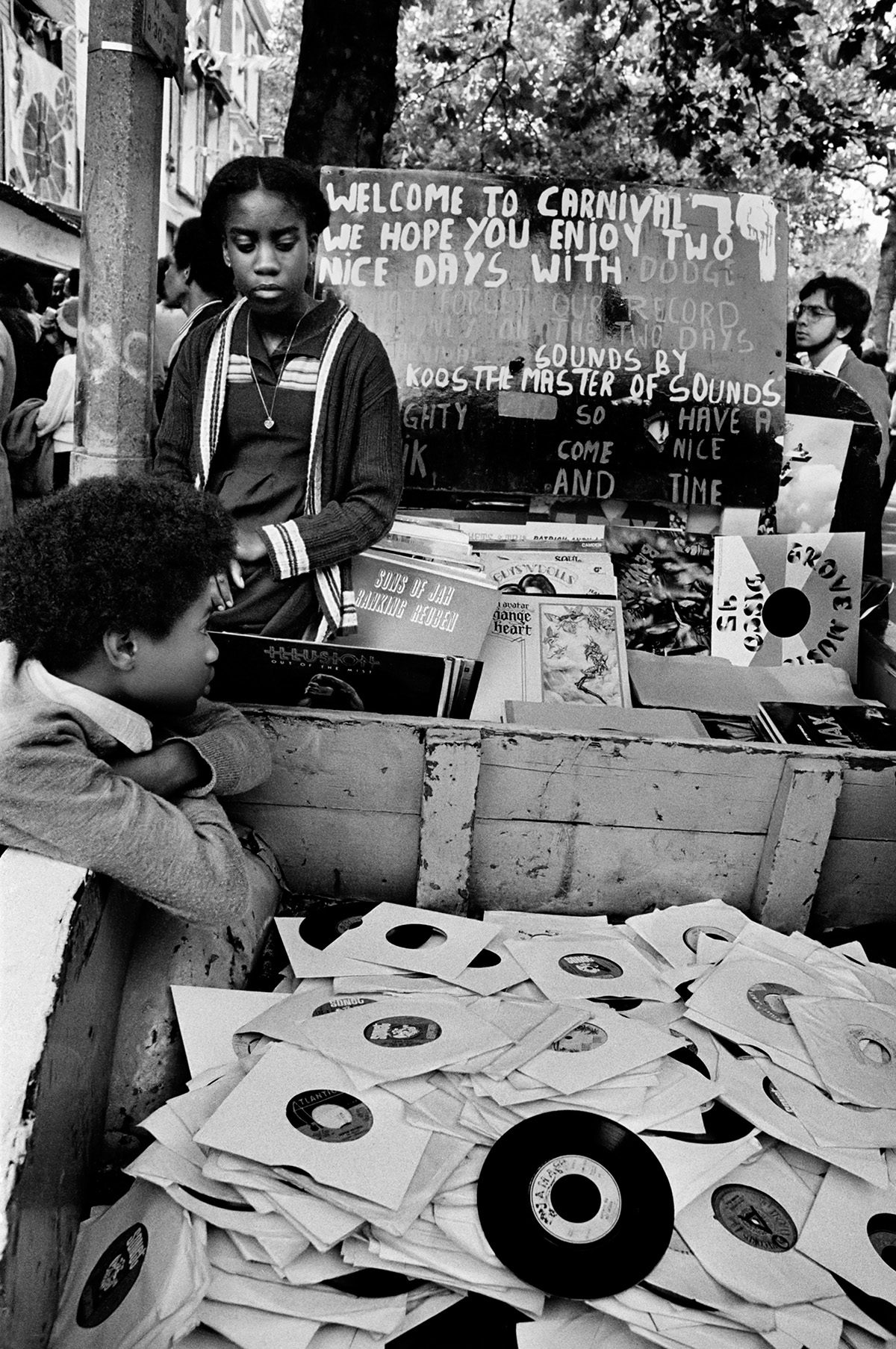 Black and white photo of two young people sat next to a large container filled with vinyl records