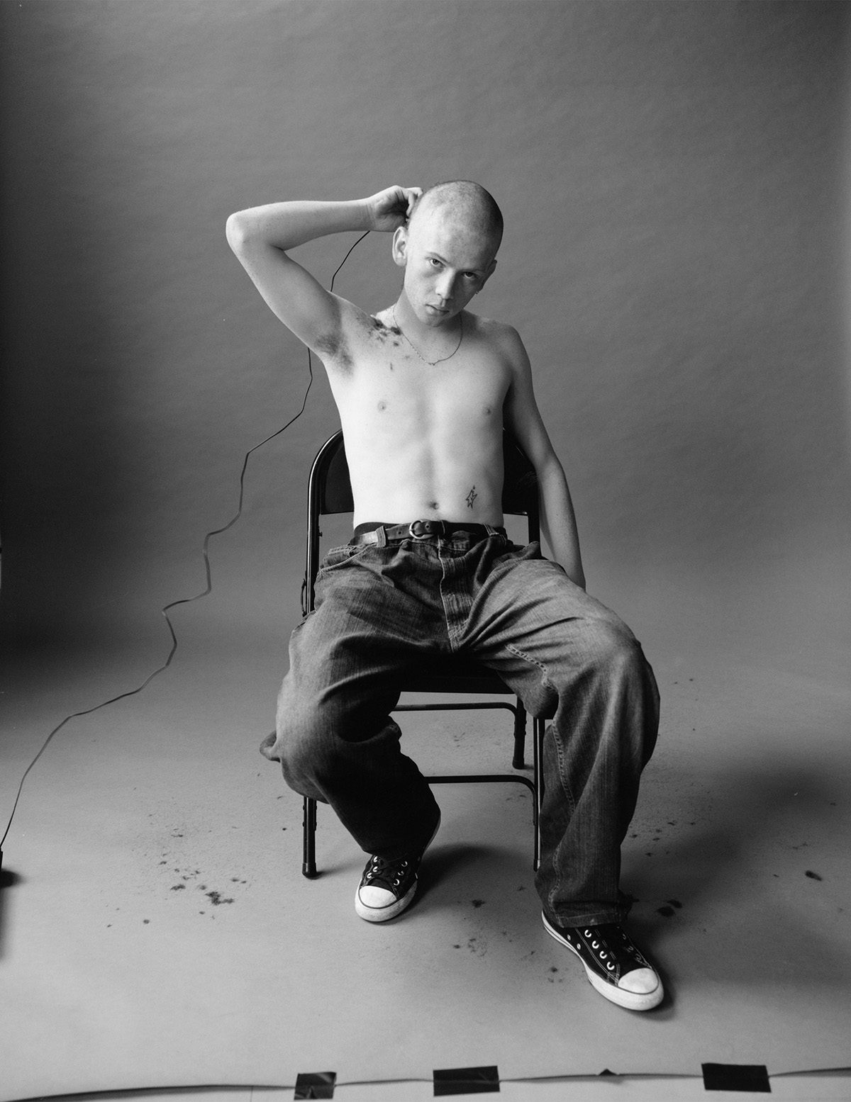 Black and white photo of a young person wearing jeans and trainers sat on a chair in a studio environment buzzing their hair