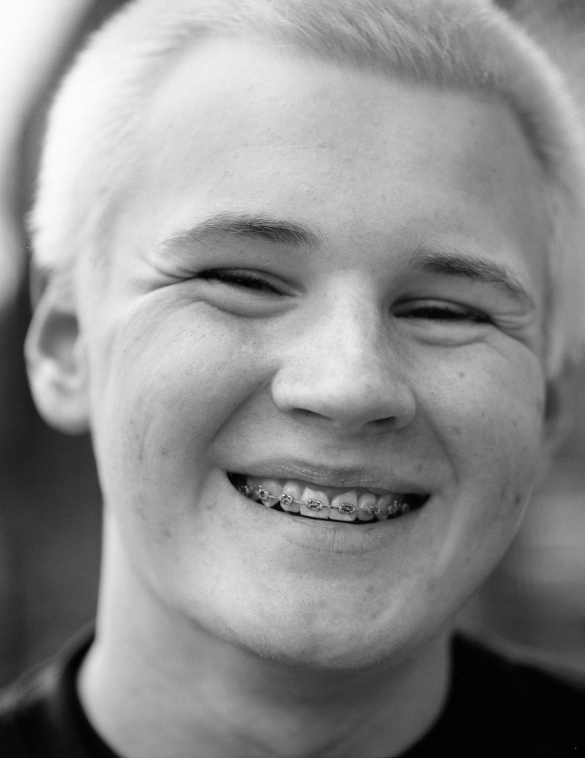 Black and white photo of a young person with light cropped hair and braces smiling at the camera
