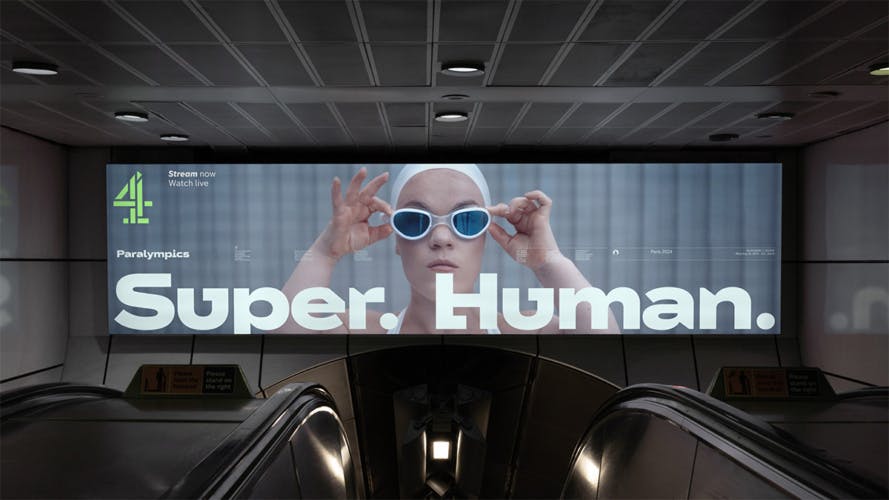 Wide outdoor advert positioned over a tube escalator entrance, featuring the slogan 'Super Human' laid over a photo of swimmer Ellie Simmonds wearing goggles.