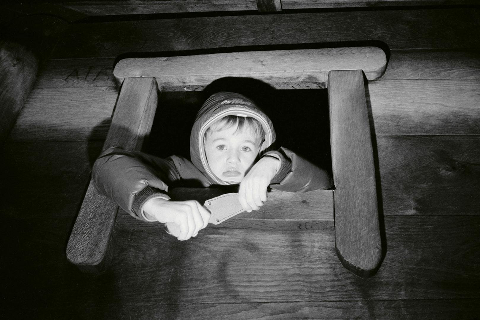 Black and white photo of a young boy wearing a hooded coat, poking out from a small window with a glum expression