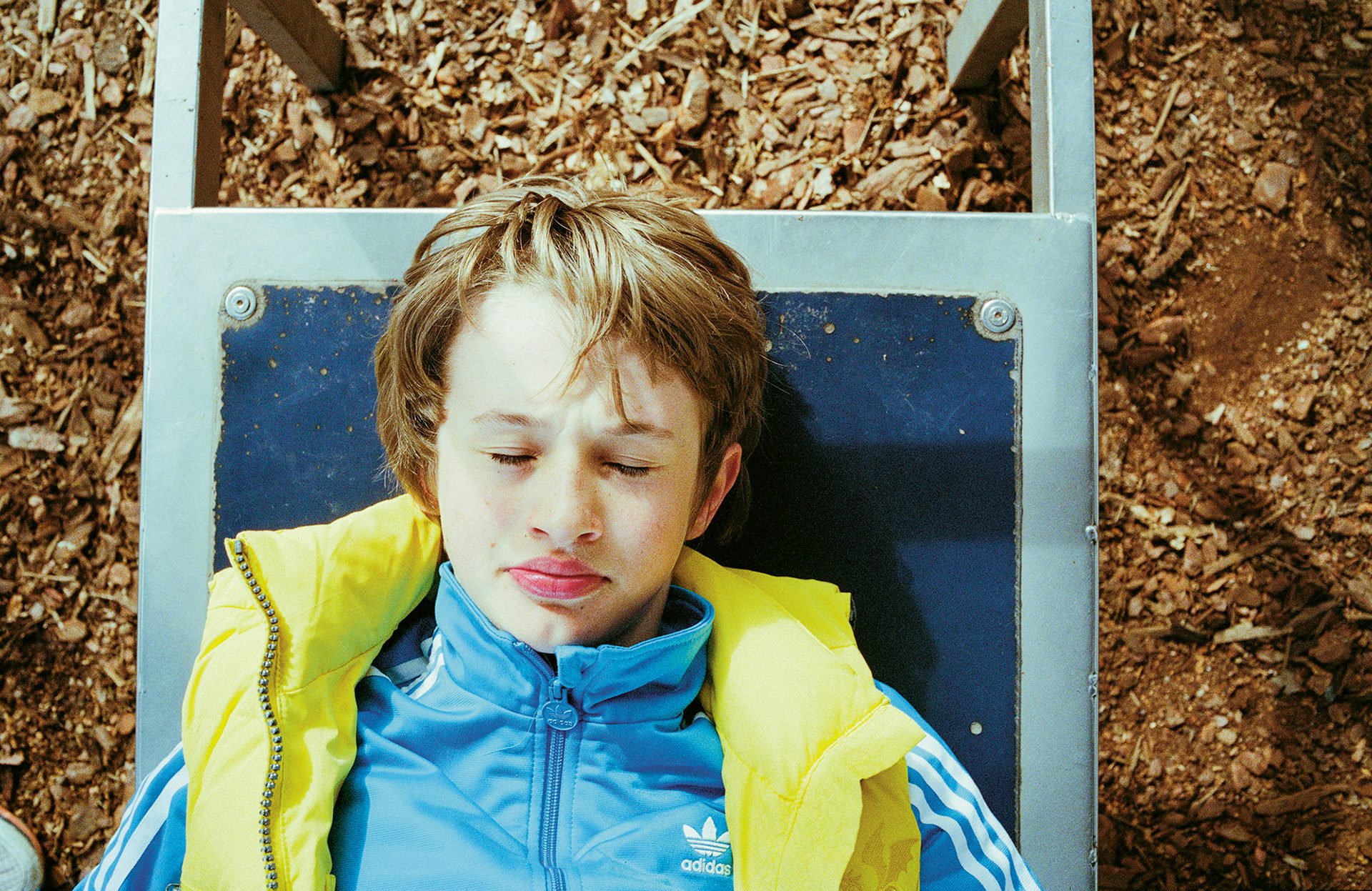 A boy with his eyes closed lying on a bench while wearing a light blue Adidas track jacket and a bright yellow vest over the top