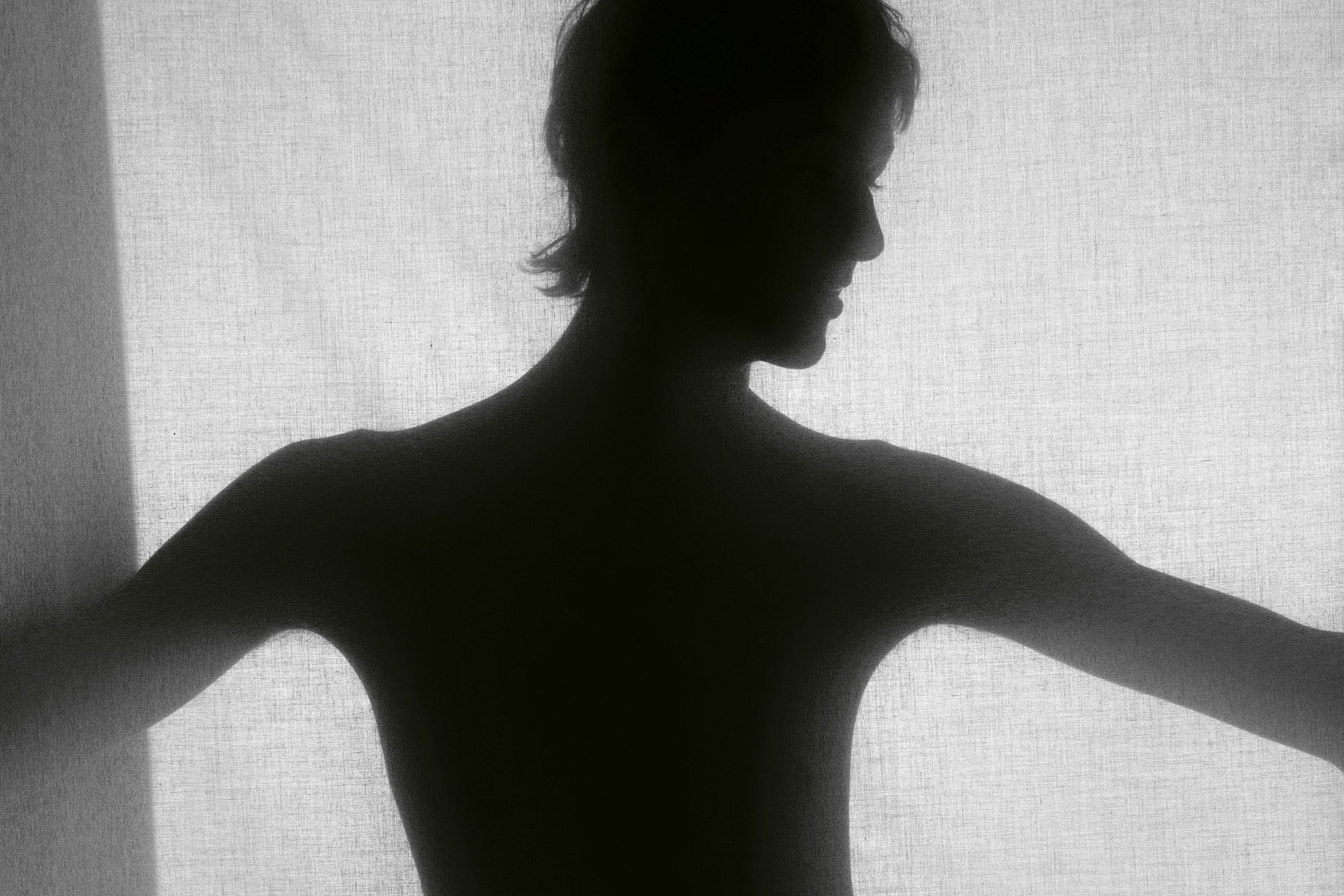 Silhouette of a boy with his arms outstretched standing in front of a piece of light fabric