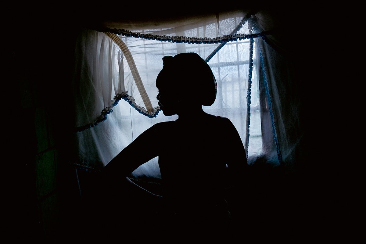 A silhouette of a person wearing a head wrap standing in front of a window covered by a thin sheer curtain
