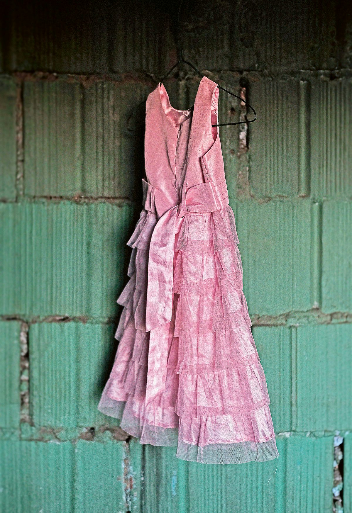 A pink tiered dress with a bow hanging on a wire hanger in front of a green wall