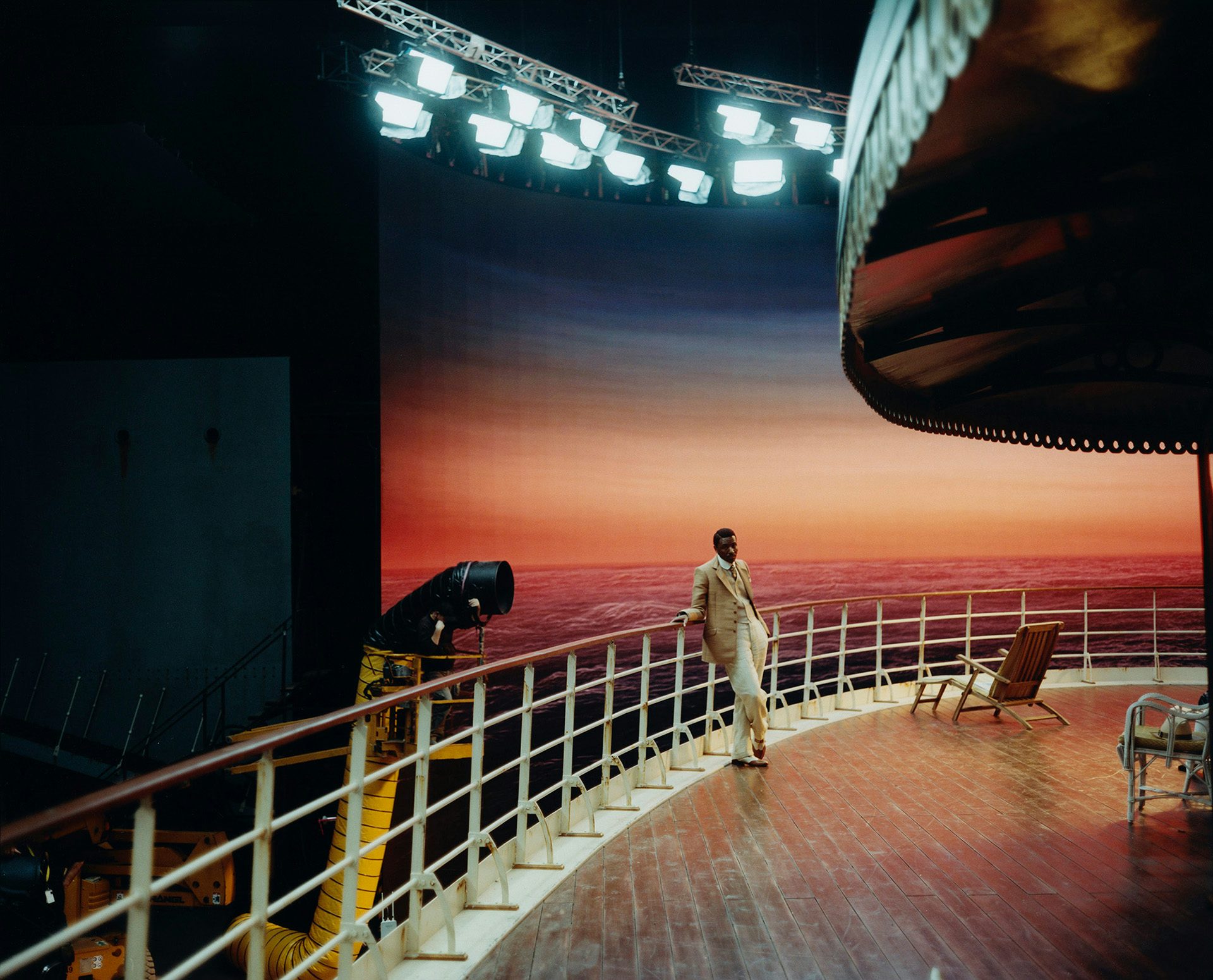 A person wearing a suit standing on a Poor Things film set of a cruise ship deck in front of a bright sunset with lighting and rigs visible