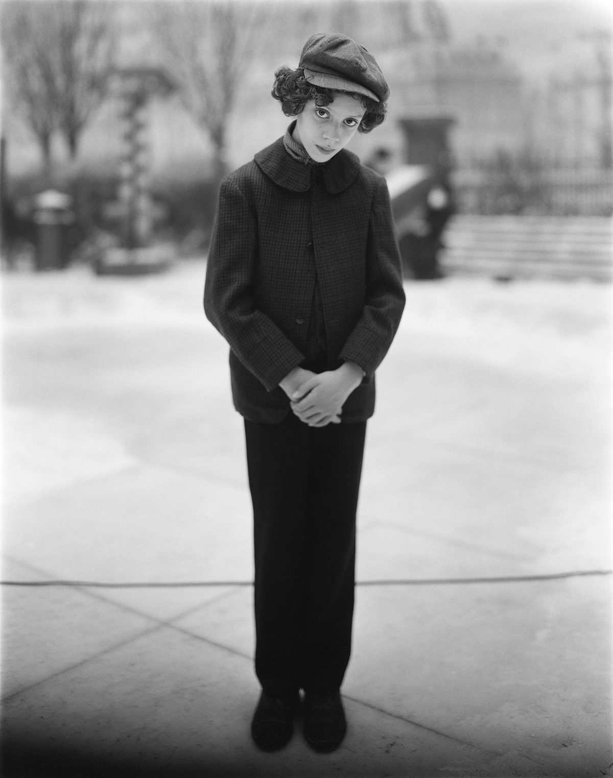 Black and white photo of a young person the set of Poor Things wearing Victorian era-style trousers, jacket and hat, with their hands crossed in front of them