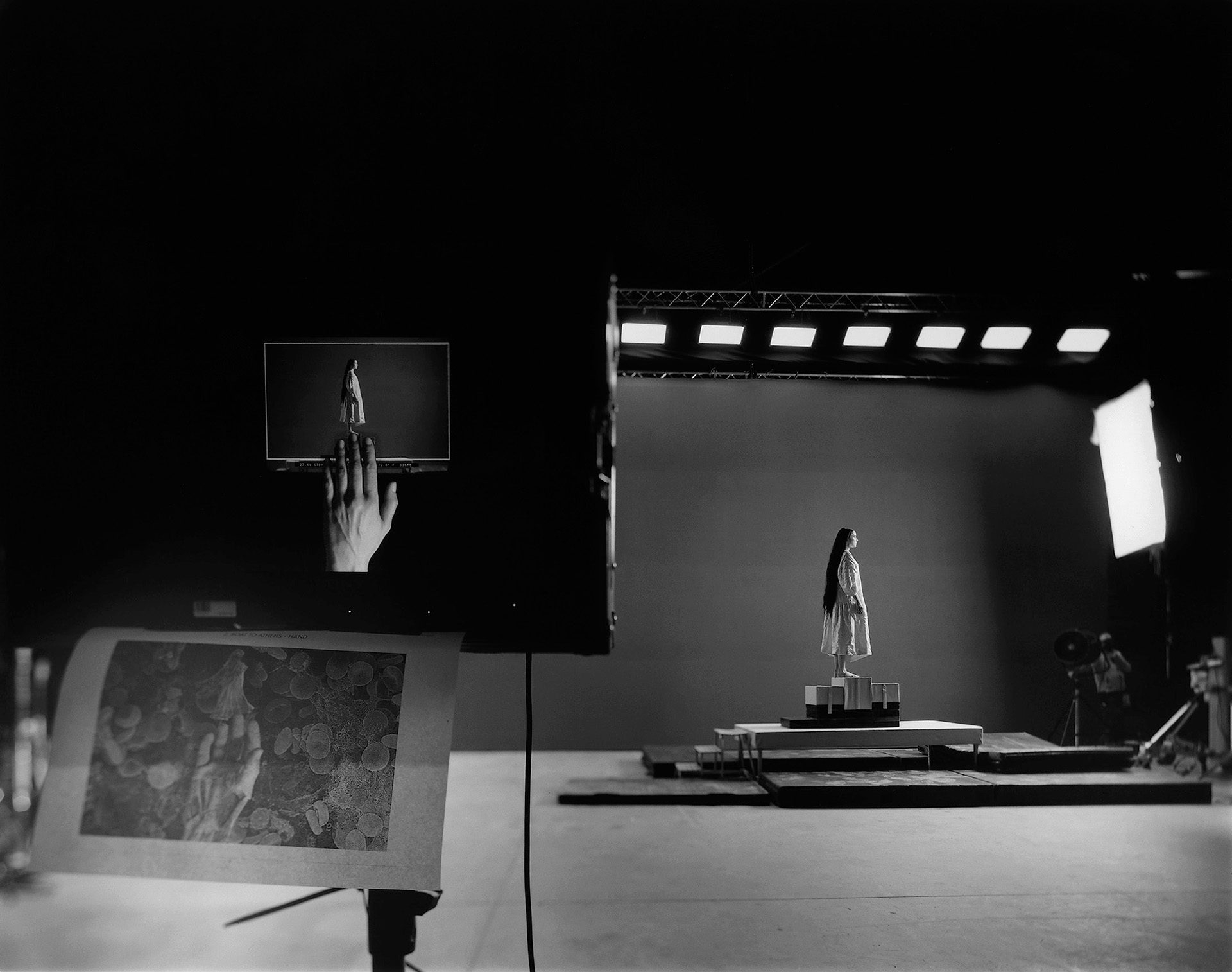 Black and white photo of Emma Stone on the set of Poor Things, standing on a podium in the middle of a large empty set, with a monitor showing the same scene visible in the foreground