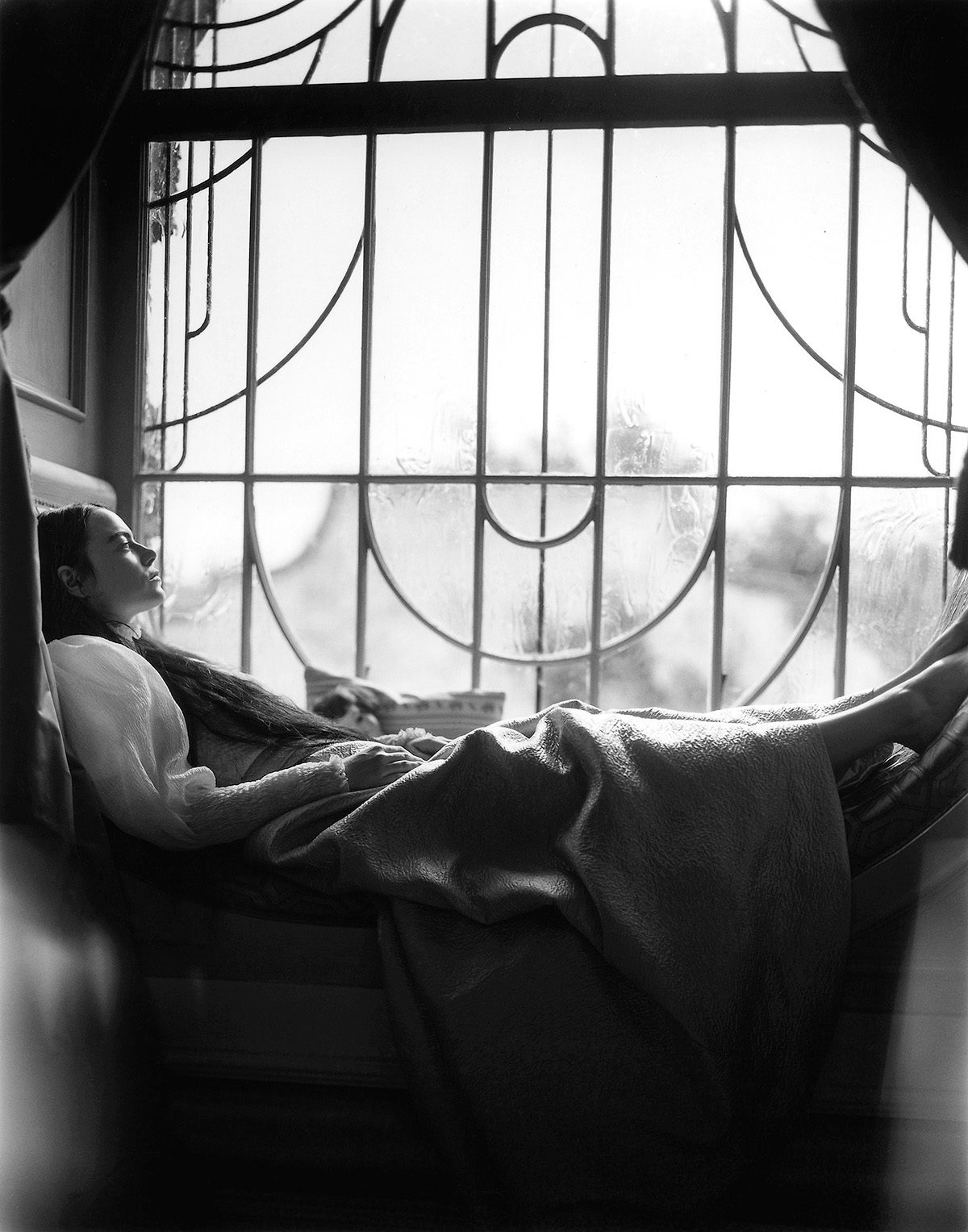Black and white photo of Emma Stone lying in a curved window frame looking out of a window with bars on it