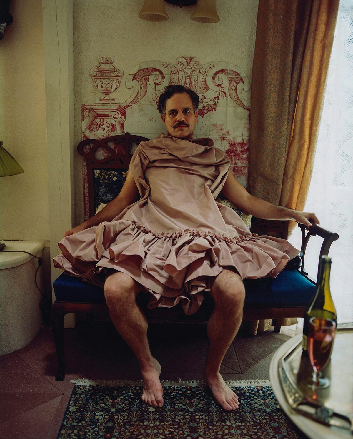 Mark Ruffalo on the set of Poor Things sat on a blue ornate chair with pink frilly fabric draped over him