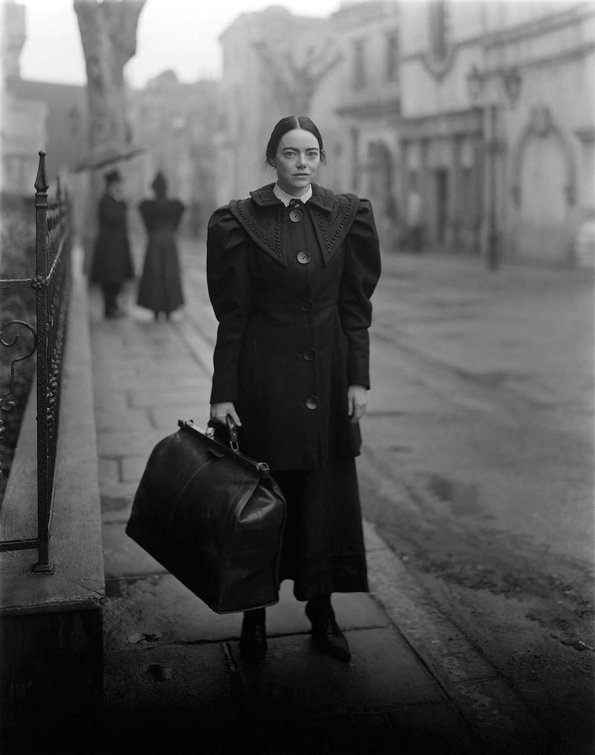 Black and white photo of Emma Stone wearing a black puff sleeved dress and suitcase in front of a Victorian era London street