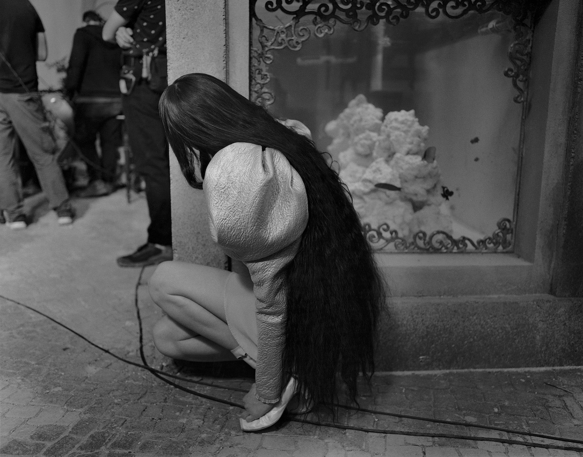 Black and white photo of Emma Stone wearing a short puff sleeved dress and long hair below her waist, crouching and looking around a wall