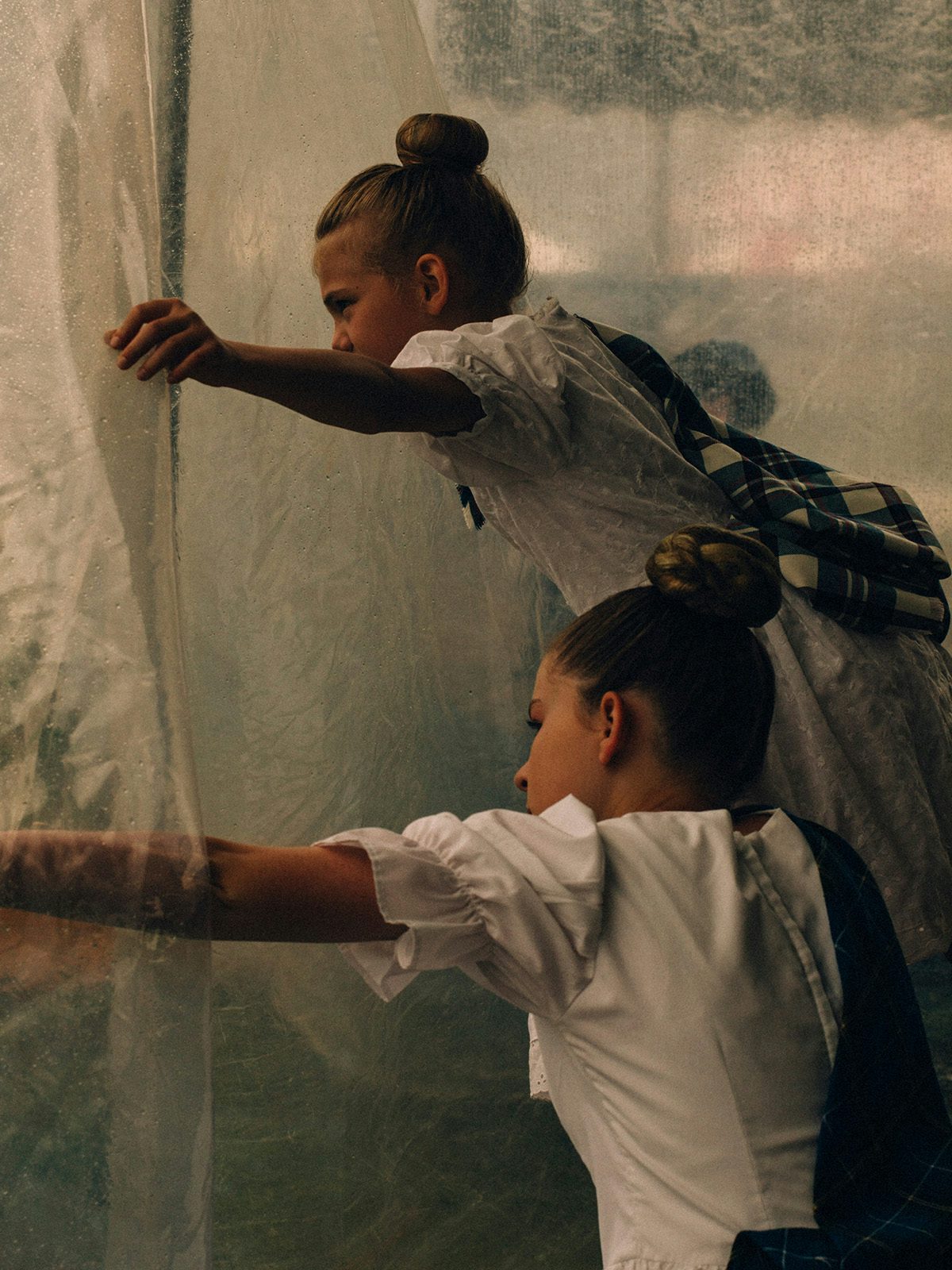 Two young girls with their hair tied in buns peering out through a sheer curtain