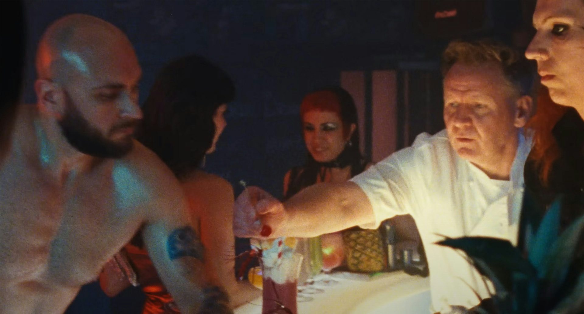 Still from the music video for Treat Each Other Right by Jamie xx, showing a topless muscular man with a beard standing at a bar opposite a Gordon Ramsay lookalike preparing a cocktail