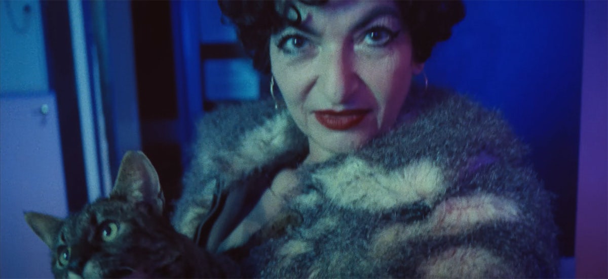 Still from the music video for Treat Each Other Right by Jamie xx, showing a woman with short hair, hoop earrings and dark red lipstick wearing a fur coat and holding a coat against her chest