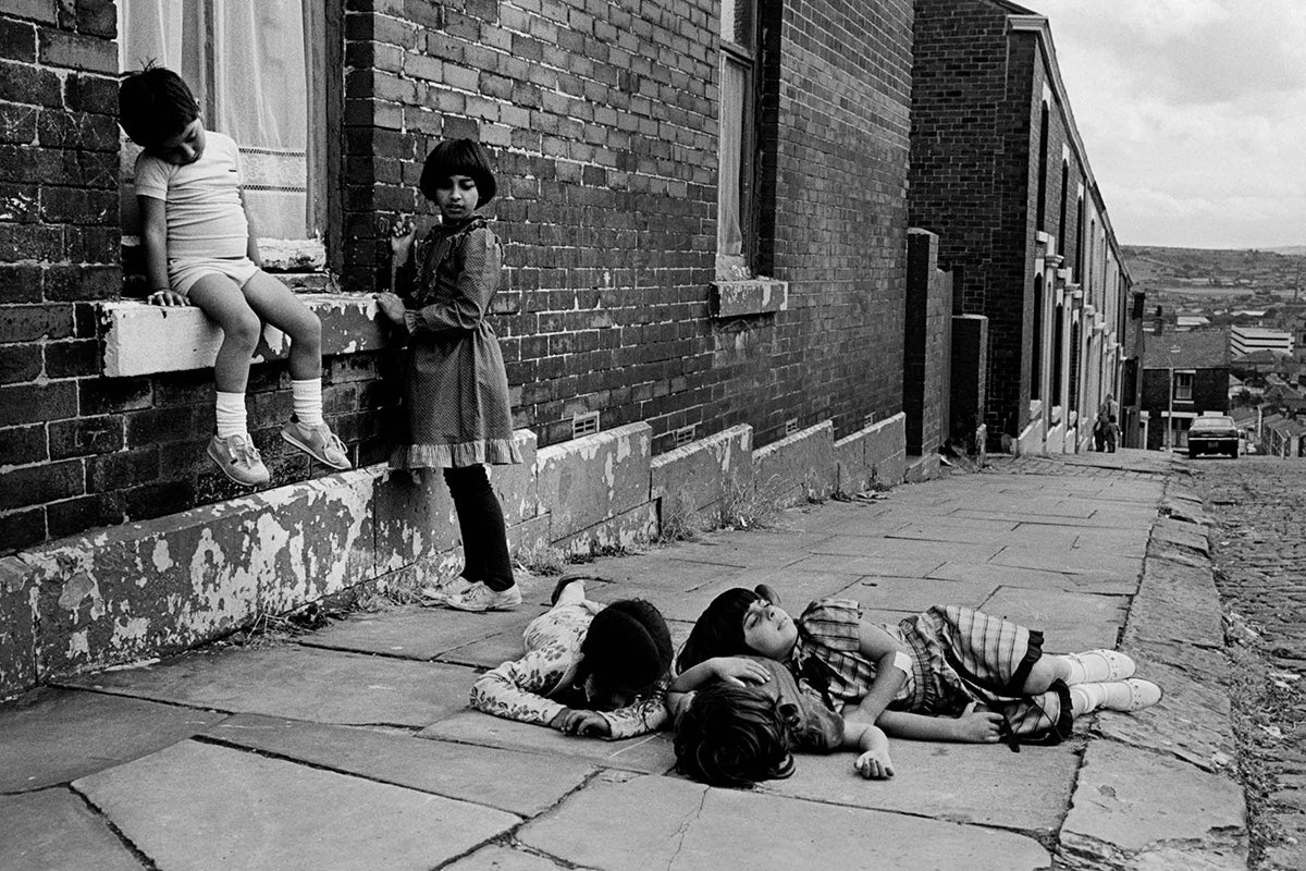 Black and white photo of three children lying down on a pavement, with two children leaning against a house, in a hilly terraced street