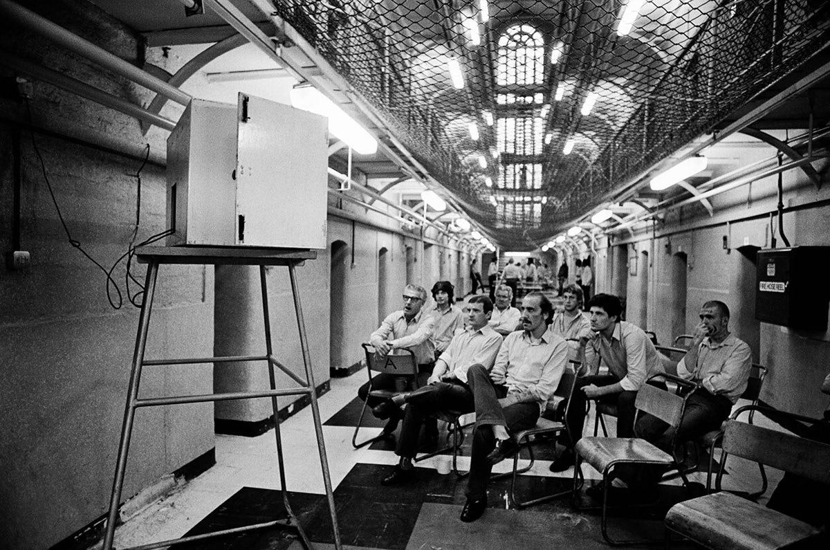 Black and white photo of a group of men sat on rows of chairs watching a television set in what appears to be a prison hall