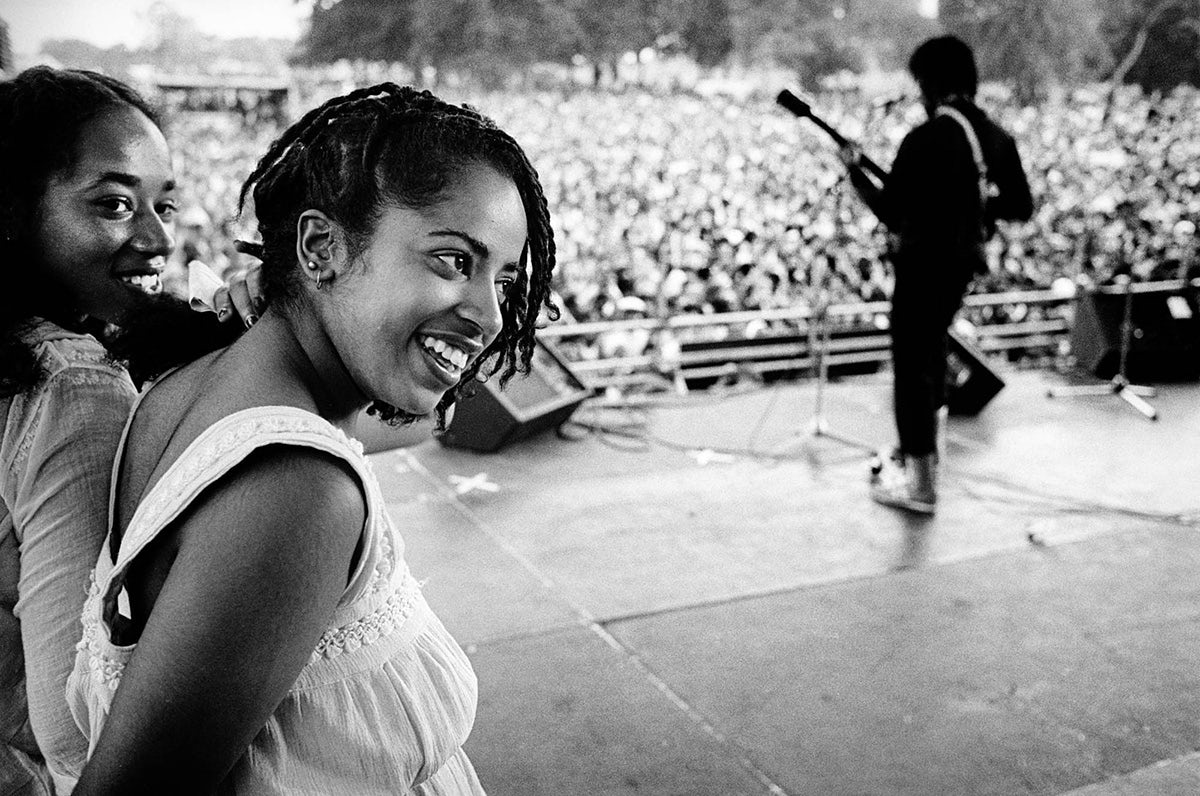 Black and white photo of a smiling young adult with braided hair standing backstage as a person at the front of the stage performs to a crowd