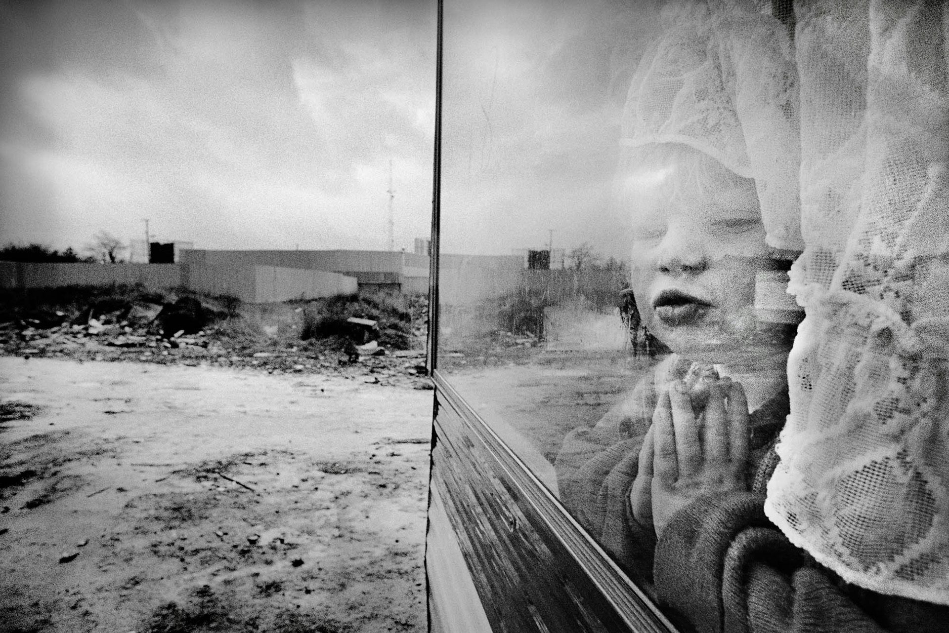 Black and white photo of a child peering underneath a net curtain through a window that looks out onto a dishevelled yard