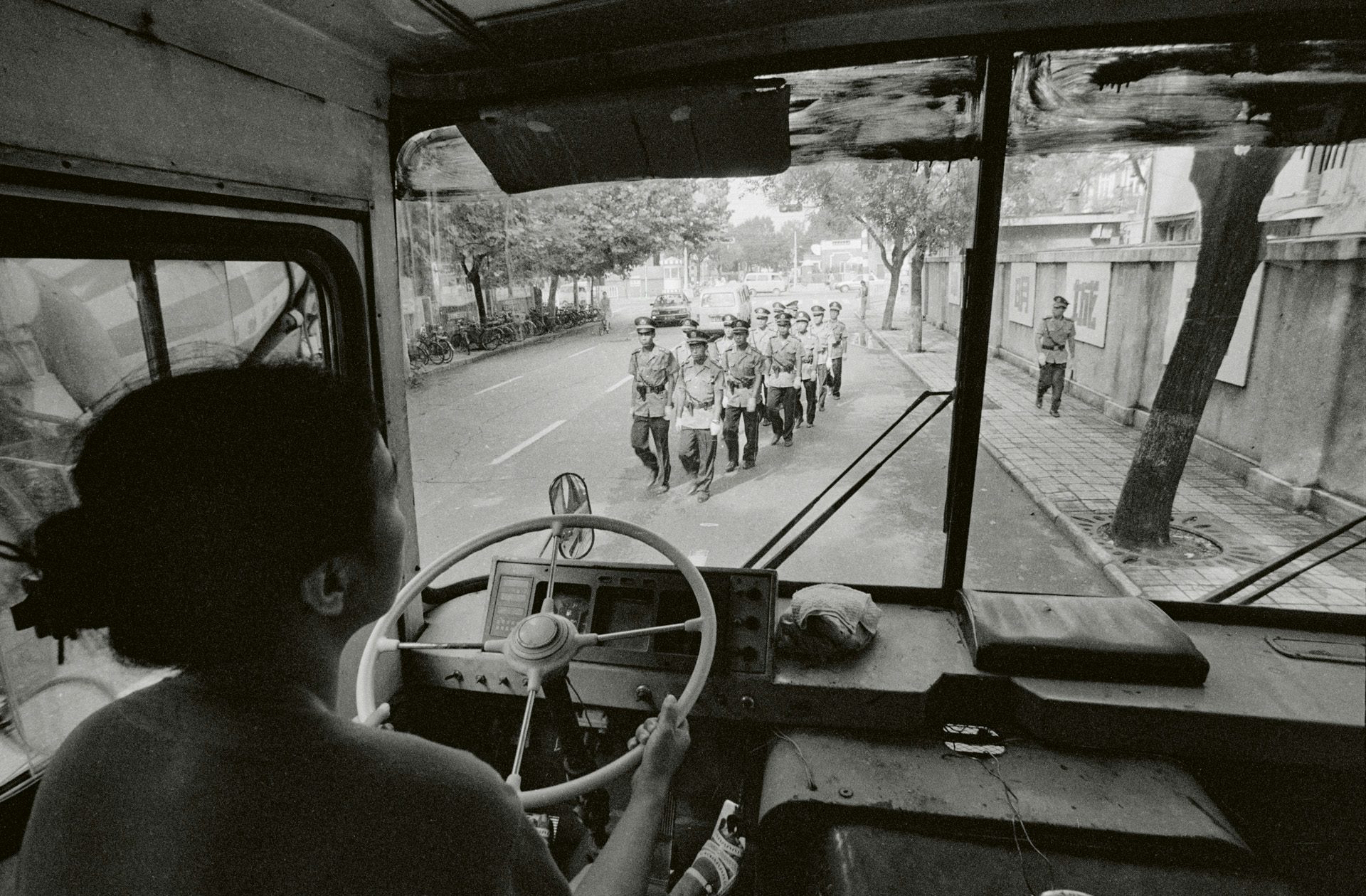 Black and white photo by Mo Yi showing armed guards marching in a street seen through a bus front window, showing the bus driver in the foreground